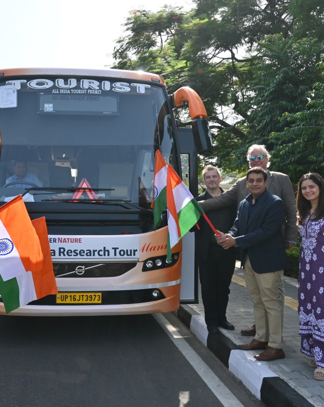 Four people holding up flags of India. Behind them is the India Research Tour bus